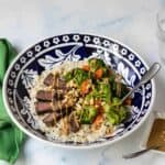 grain bowl with steak, brown rice, broccoli, carrots, and peanut sauce in blue bowl with green napkin