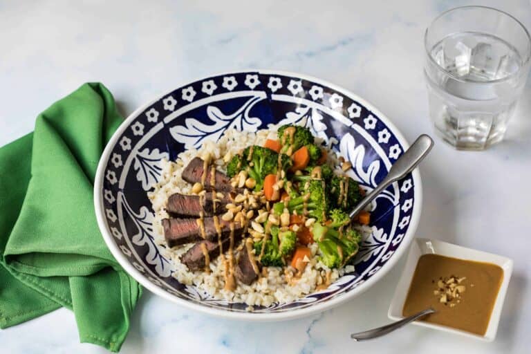 grain bowl with steak, brown rice, broccoli, carrots, and peanut sauce in blue bowl with green napkin