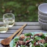 fig salad with blue cheese, pecans, and proscuitto on wooden table outside with glasses of wine, bowls, and serving utensils