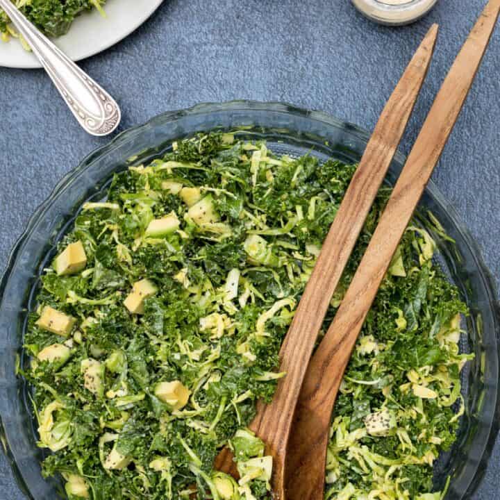 kale and brussels sprouts salad in large bowl with tongs, dressing, and small salad plate
