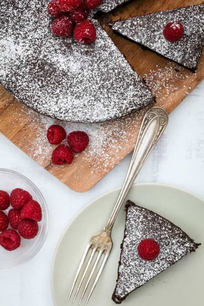almond flour chocolate cake slice on plate with full cake on cutting board behind, with raspberries