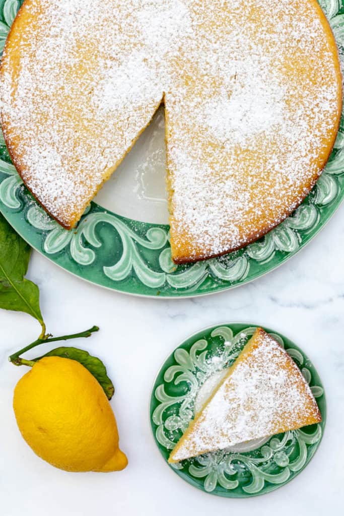 lemon ricotta cake on large plate with slice on smaller plate and lemon next to it 