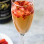close up of glass of strawberry mimosa with bowl of chopped strawberries and champagne bottle in background