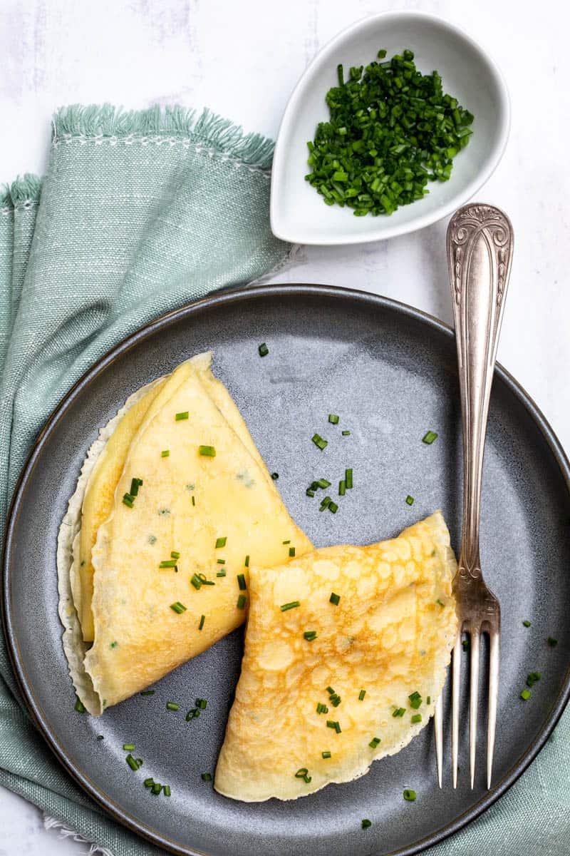 overhead shot of French crepes with eggs and cheddar  on a plate with fork and bowl of chives and napkin