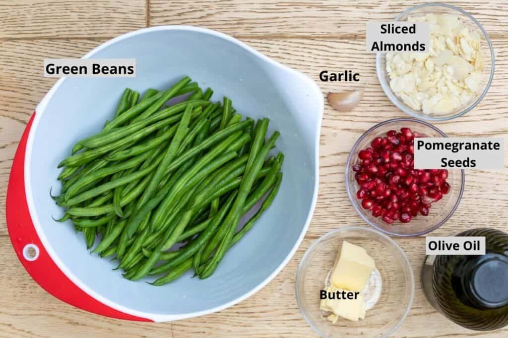 green beans in bowl with butter, pomegranate seeds, garlic, sliced almonds, and olive oil in other bowls or containers