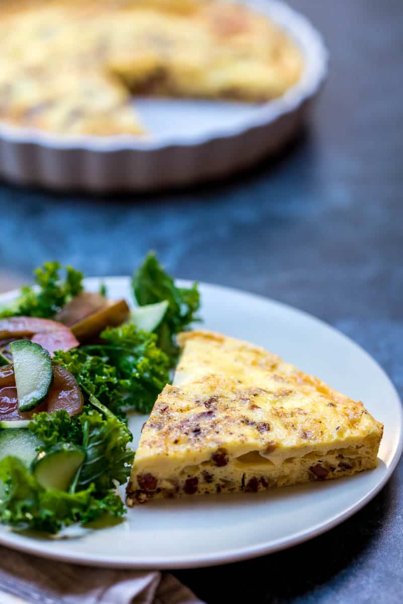 close up of slice of crustless quiche lorraine on plate next to salad with quiche pan behind it