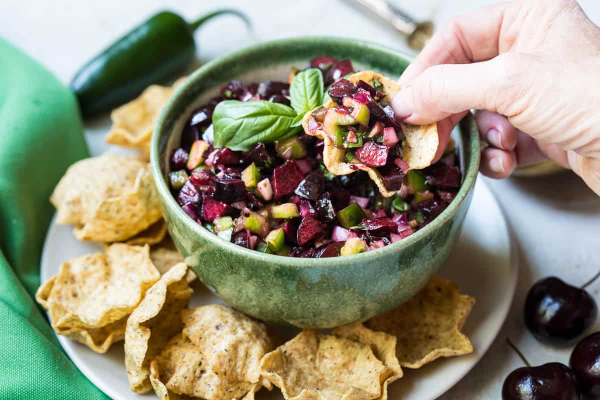bowl of cherry salsa next to tortilla chips with hand reaching in to dip a chip.