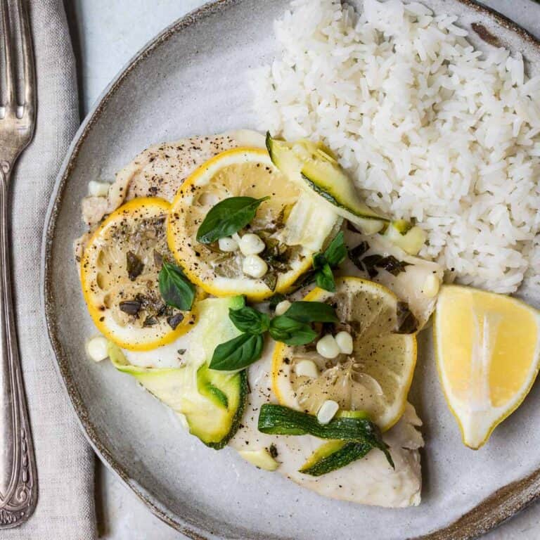 overhead view of steamed tilapia with zucchini, corn, basil, and lemon next to rice on plate.