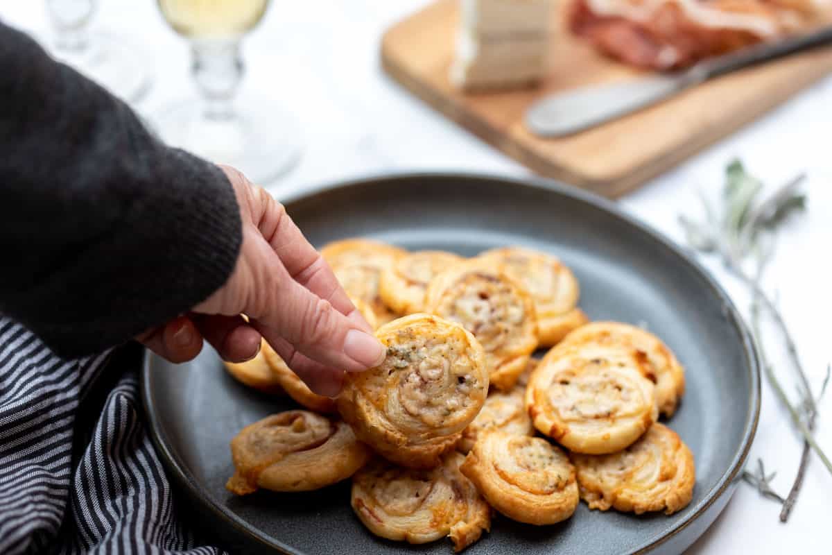 hand reaching in to grab a puff pastry prosciutto pinwheel off a plate of stacked pinwheels.
