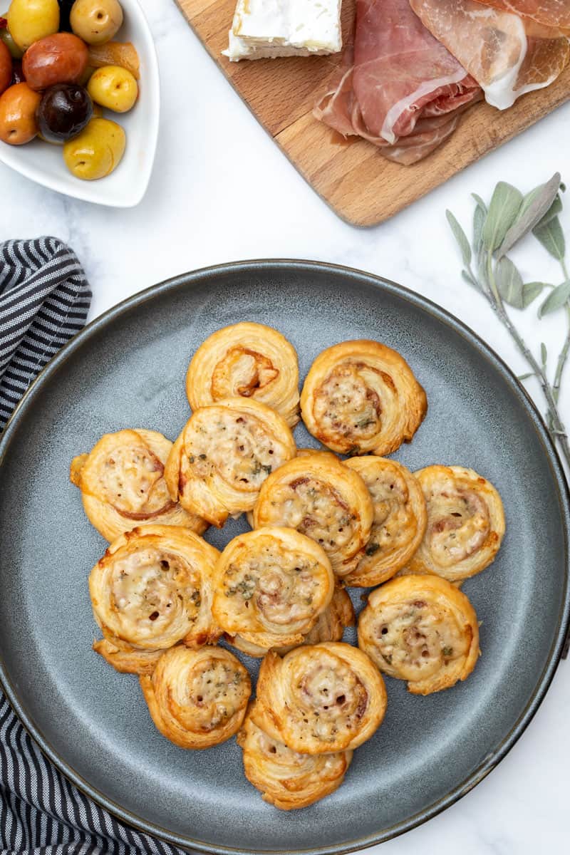 plate of puff pastry pinwheels with napkin, bowl of olives, and cutting board in the background