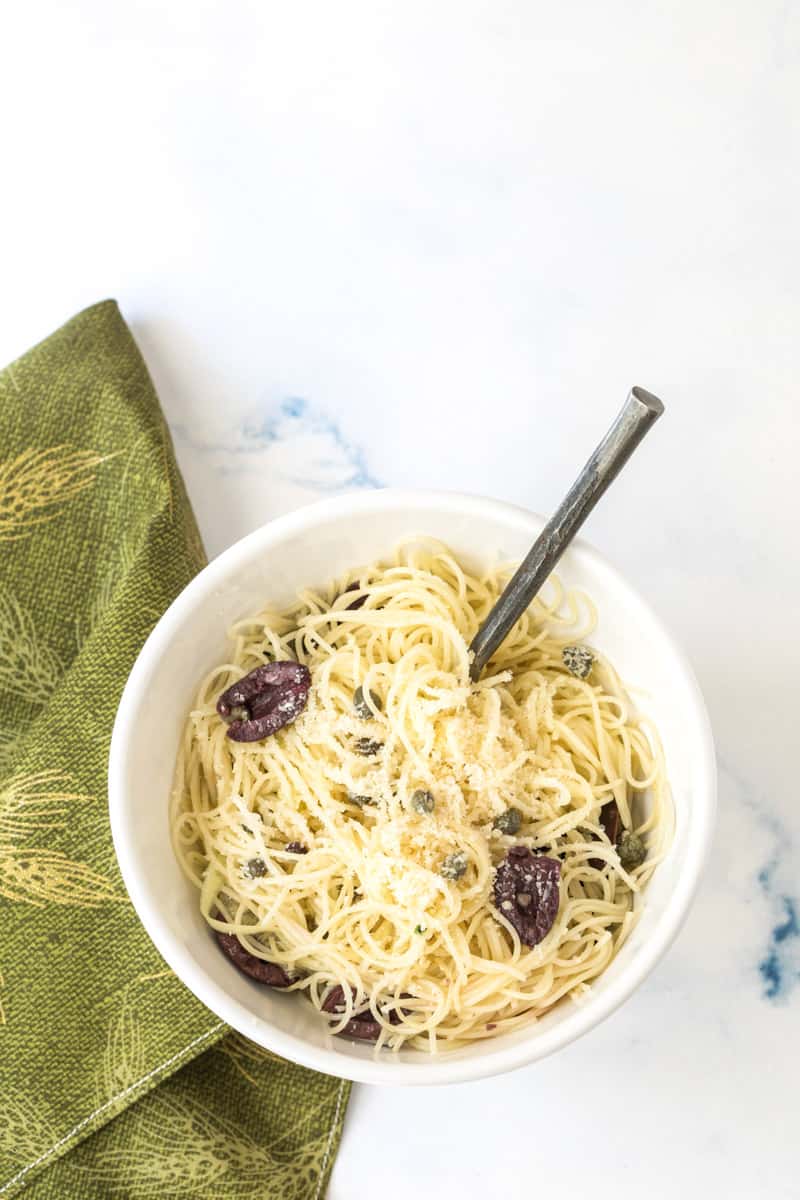 angel hair pasta with olives and capers in bowl with spoon and napkin
