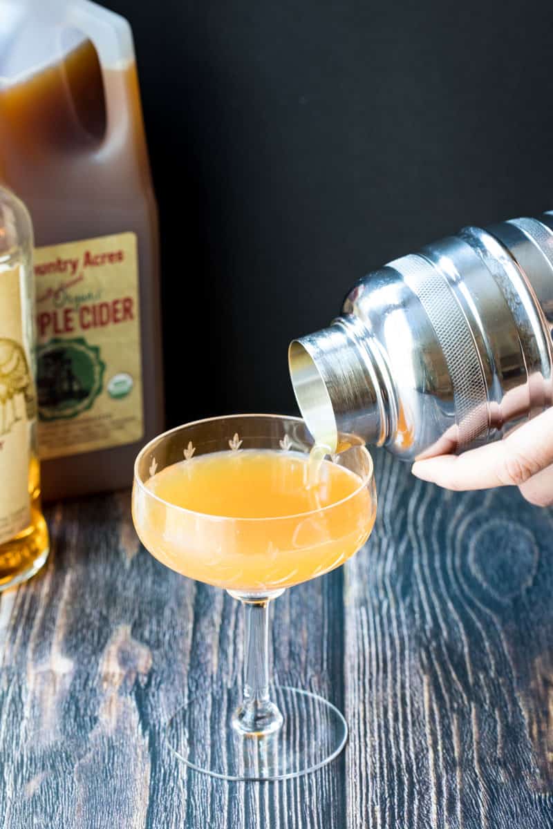 pouring apple cider bourbon cocktail from shaker into glass, next to bottle of apple cider