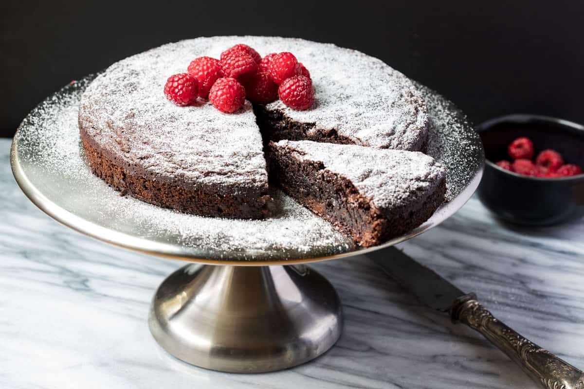 almond flour chocolate cake on cake platter with bowl of raspberries and cake knife in background