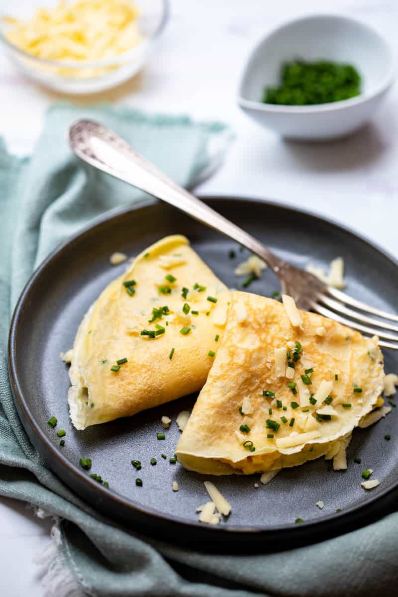plate with egg filled French crepes with fork, napkin in front of bowls of chives and cheese