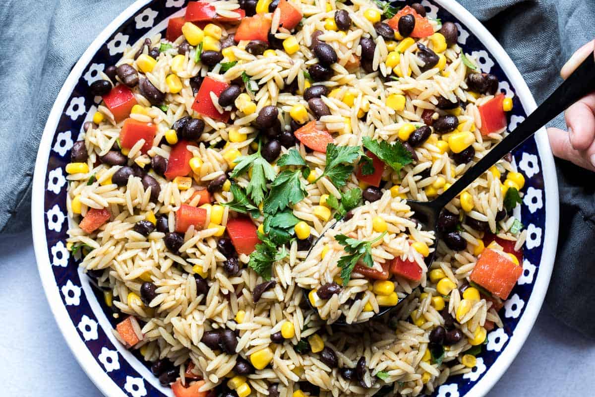 large bowl of orzo pasta salad with black beans and corn with fingers holding spoon and napkin in background