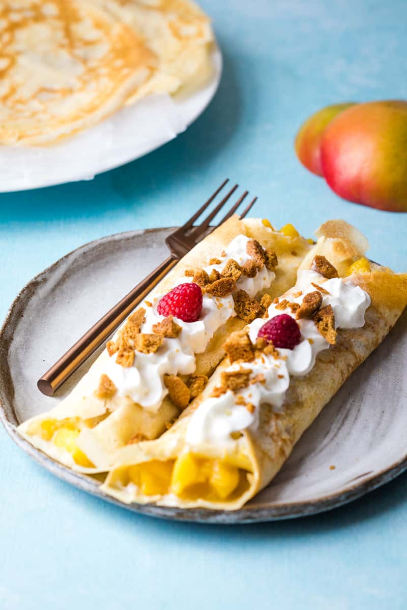 close up of two filled mango crepes topped with gingersnaps, whipped cream, and raspberries on plate with fork in front of plate of crepes and mango peels