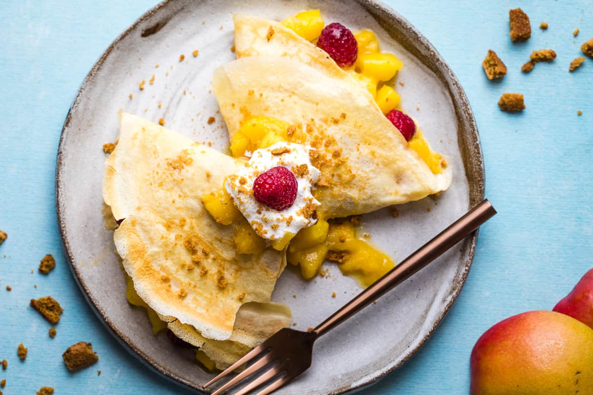 two mango crepes folded in triangles topped with mango filling, whipped cream, and raspberries on plate with fork next to mango peels and gingersnap crumbs