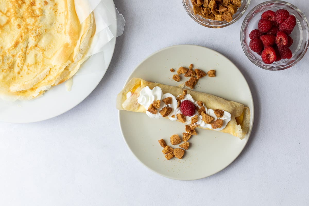 mango crepe after filling and rolling, topped by whipped cream, raspberry, and gingersnaps next to bowls of filling and plate of crepes