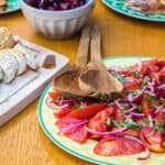 plate of tomato salad with serving utensils next to cutting board with baguette slices