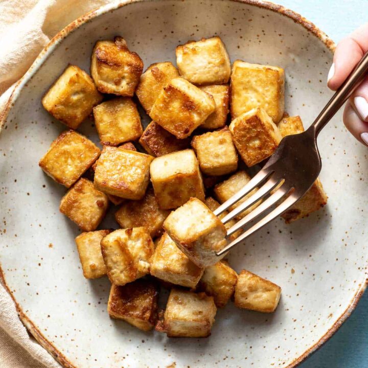 close up of shallow bowl with crispy tofu cubes and hand holding fork with one tofu cube.