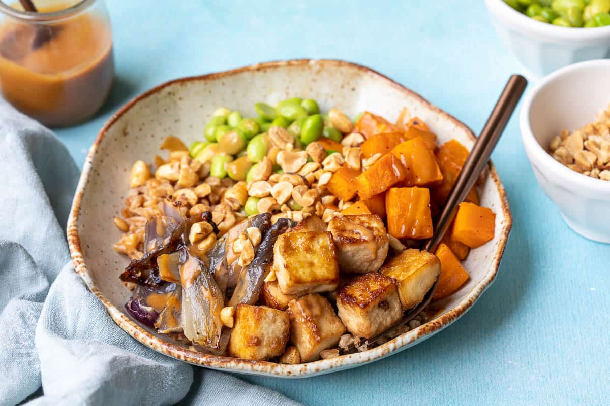crispy tofu buddha bowl with fork next to napkin, jar of peanut sauce, and bowls of edamame and peanuts.