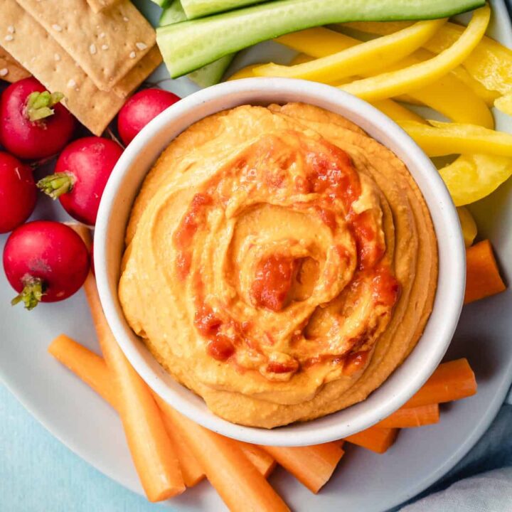 close up of harissa hummus in small bowl next to radishes, carrot sticks, and peppers.