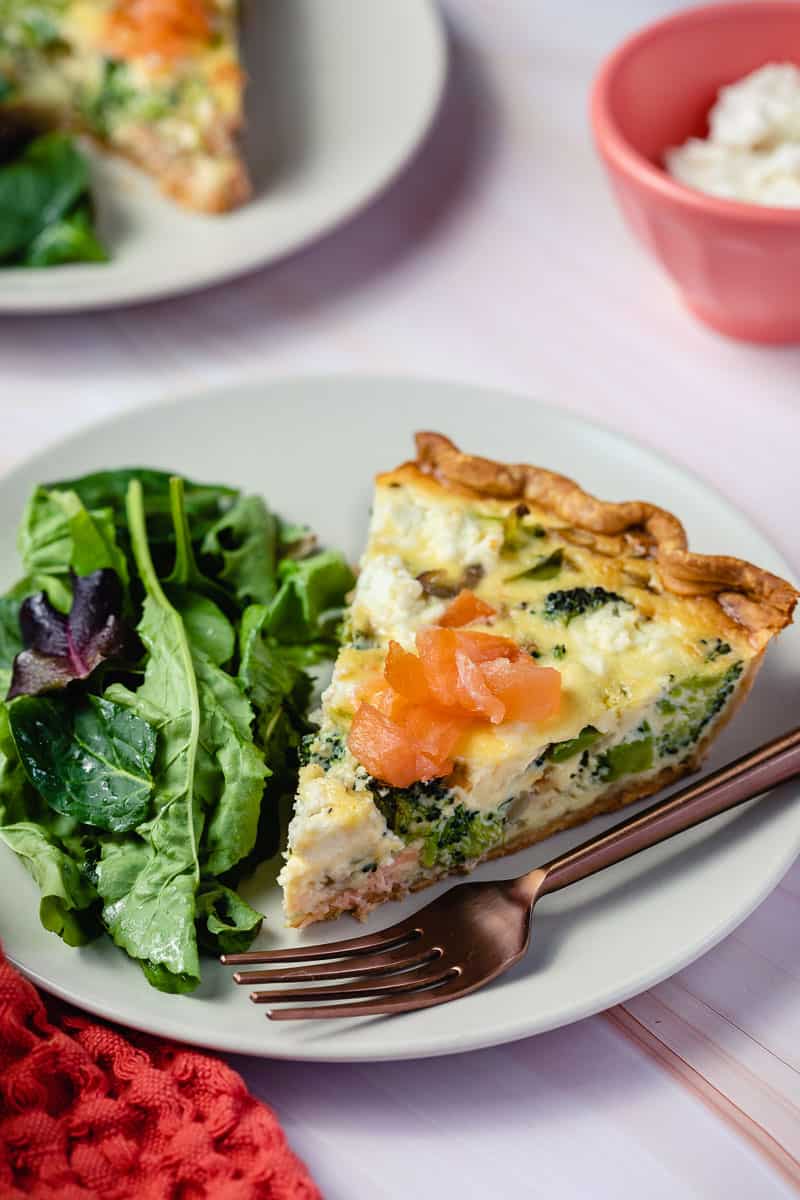 slice of salmon broccoli quiche next to salad greens and fork in front of other plate and bowl of goat cheese.