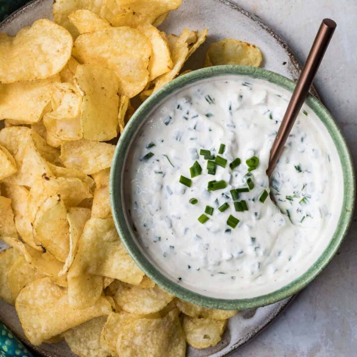 Close up of bowl of sour cream and chive dip with spoon on plate with potato chips.