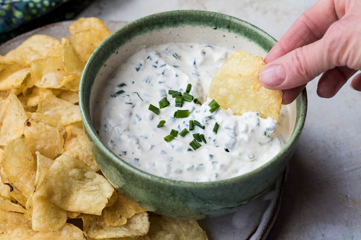 bowl of sour cream and chive dip with hand reaching in to dip potato chip.