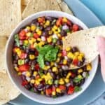 overhead shot of bowl of black bean and corn salsa with chip being dipped on serving platter next to tortilla chips and kitchen towel.
