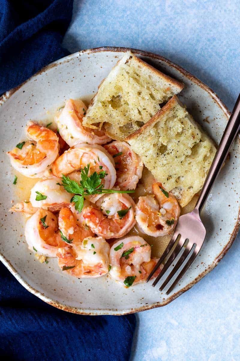 plate with shrimp scampi without wine next to garlic bread and fork, with blue napkin.