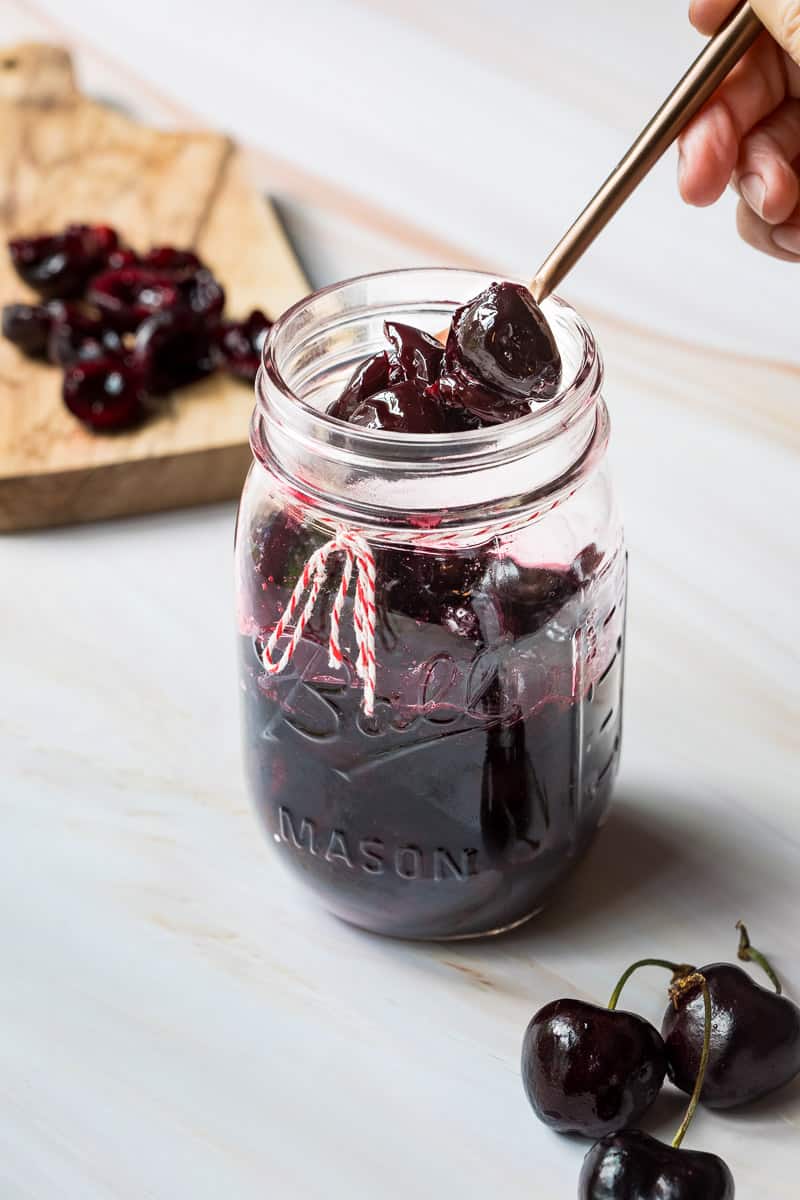 jar with homemade cherry compote with spoon in front of cutting board with cherry halves and behind a few cherries.