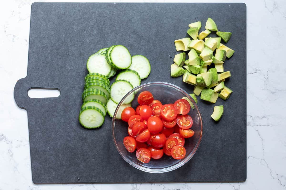 sliced cucumbers, cubed avocado chunks, and halved cherry tomatoes on cutting board.