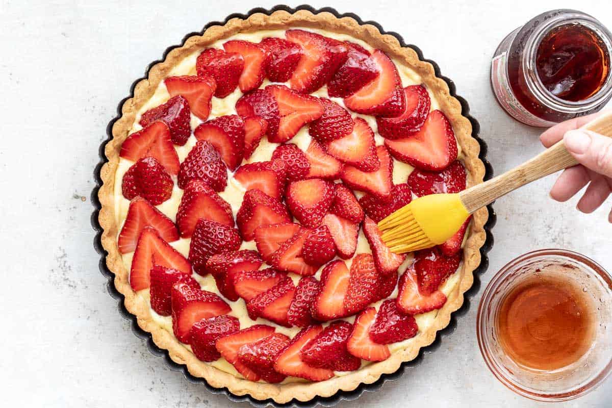 hand holding pastry brush brushing strawberry glaze on tarte aux fraises, next to small bowl of jam and jam jar.