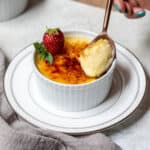 ramekin of vanilla bean creme brûlée with strawberry and mint leaves and spoon scooping out custard.