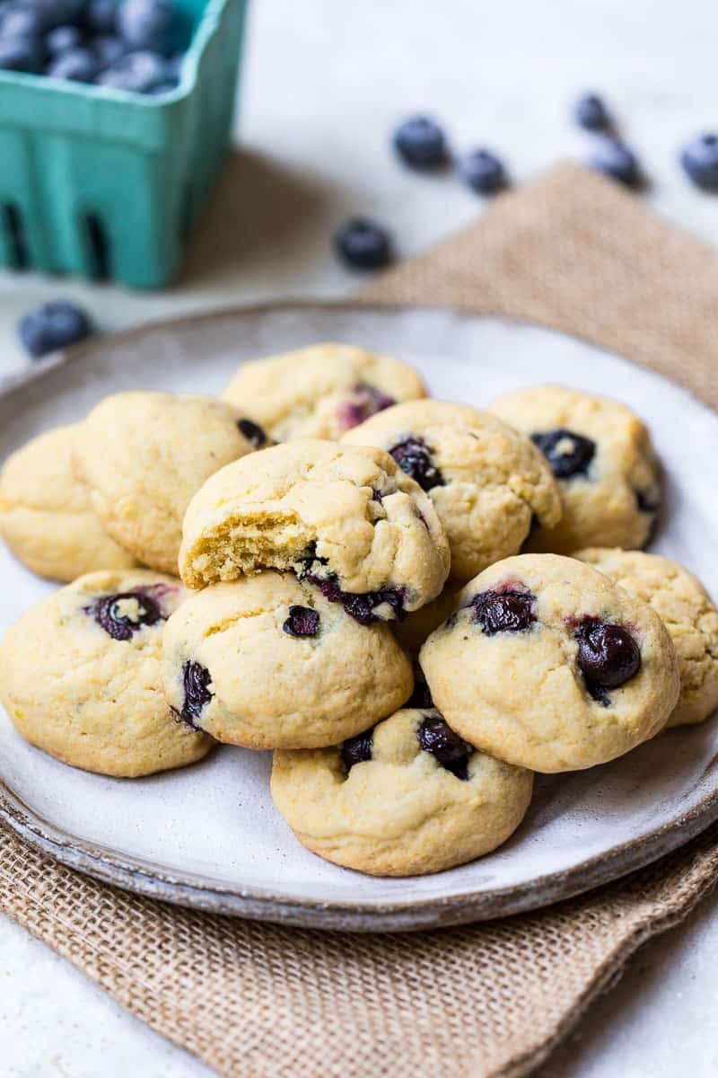 stack of lemon blueberry cookies on plate on top of burlap in front of basket with blueberries.