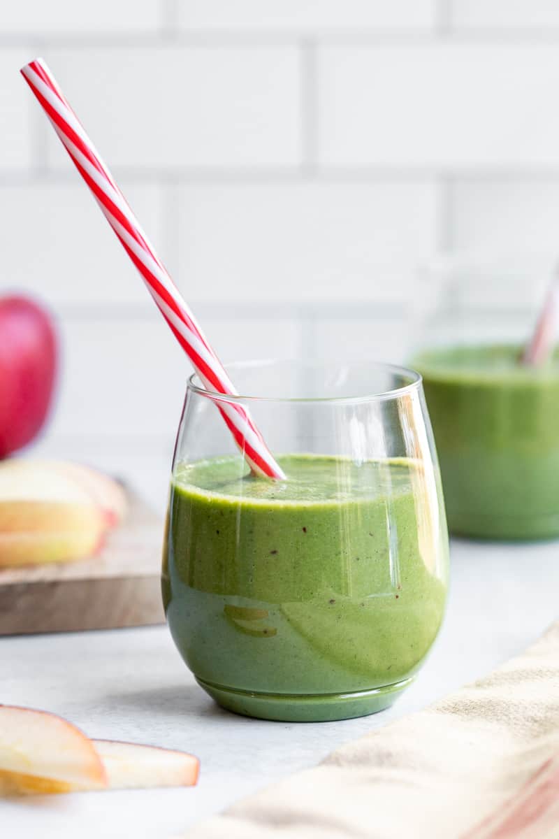 glass with apple spinach smoothie with red and white straw in front of cutting board with apple slices and other green smoothie.