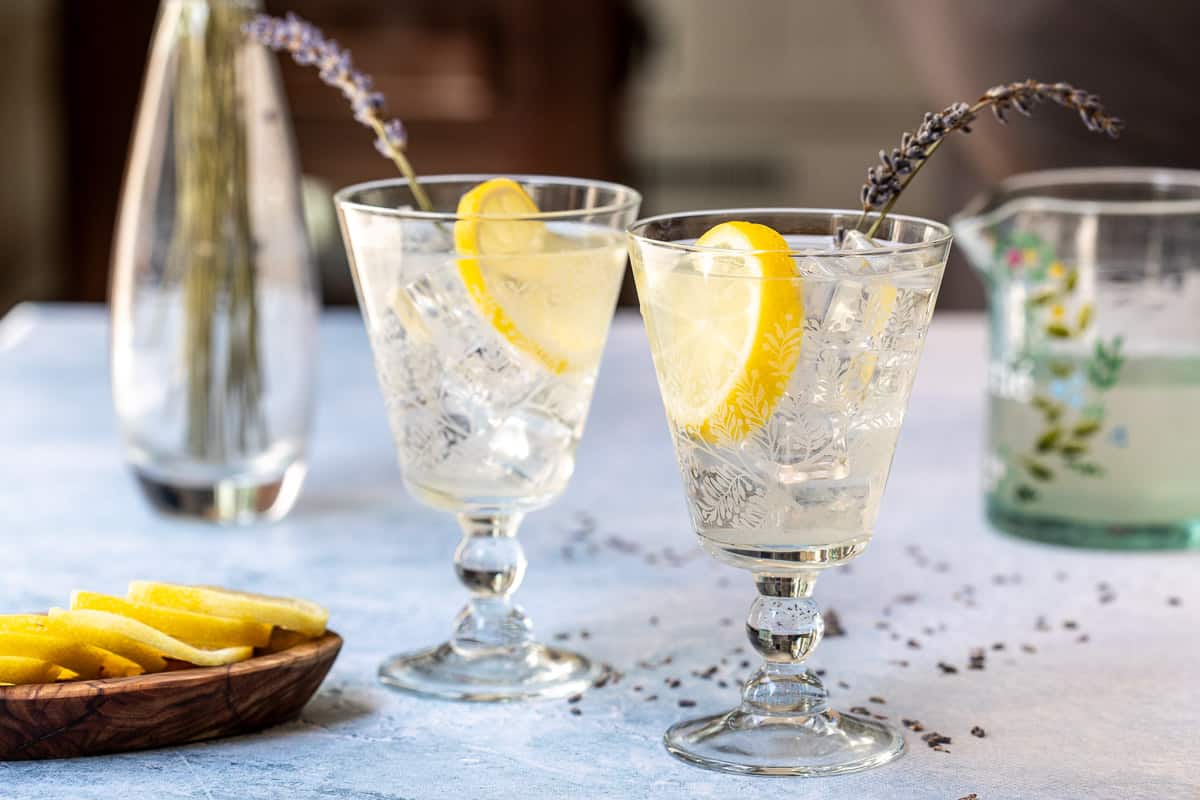 two glasses of lavender lemonade next to lemon slices, pitcher, and vase with lavender.