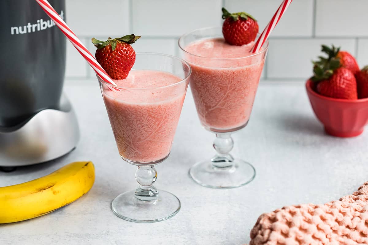 two glasses with dairy-free banana strawberry smoothie next to banana, blender, and bowl of strawberries.