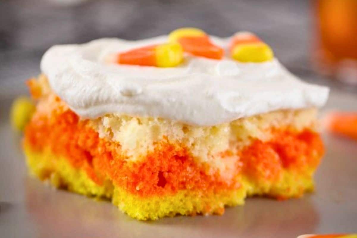 A slice of layered yellow, orange, and white cake with whipped topping and candy corn pieces on top.