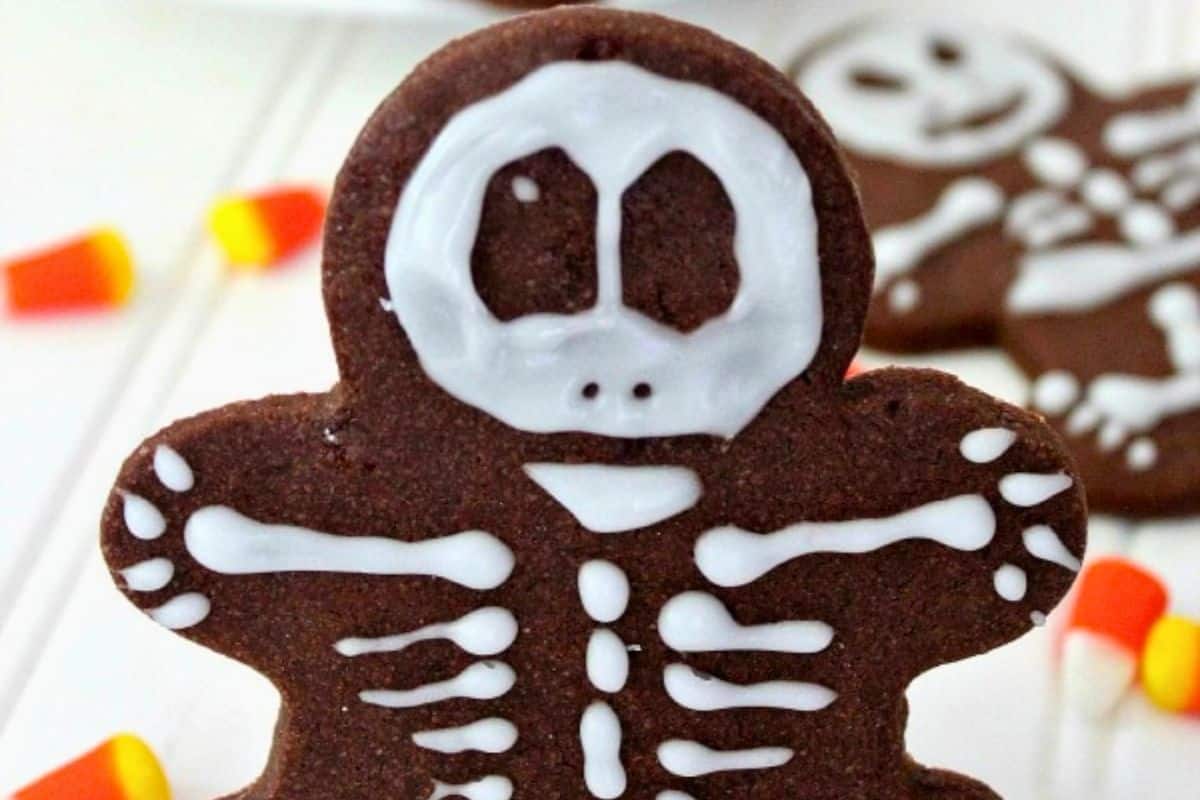 Chocolate skeleton-shaped cookie decorated with white icing to resemble bones, with pieces of candy corn in the background.