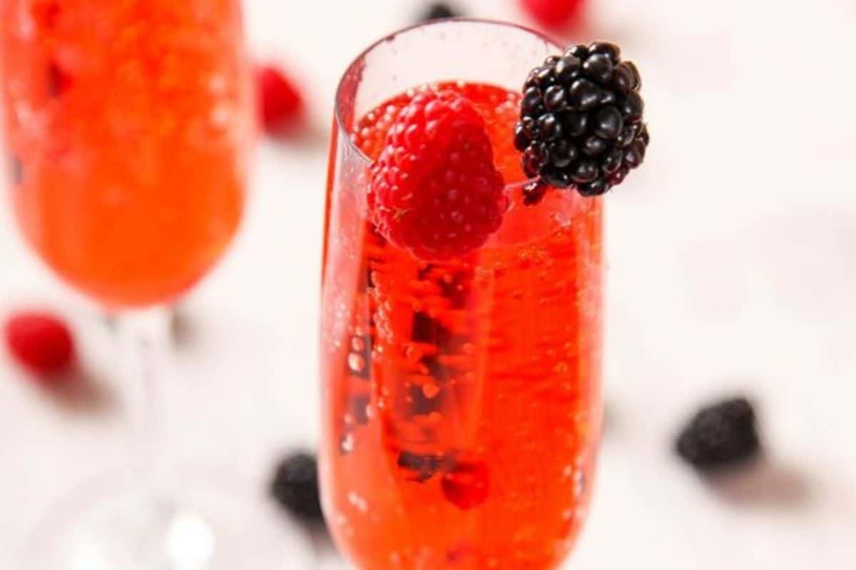 A glass of red sparkling drink garnished with a raspberry and a blackberry, with another similar glass and scattered berries in the background—perfect for those seeking festive cocktails to brighten up any celebration.