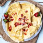 stack of French crêpes on a plate topped with chopped strawberries and next to whole strawberries.