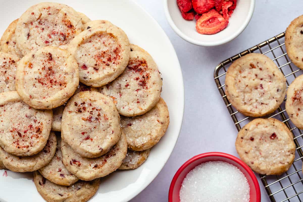 strawberry shortbread cookies with bowl of sparkling sugar and bowl of dried strawberries next to cooling rack.
