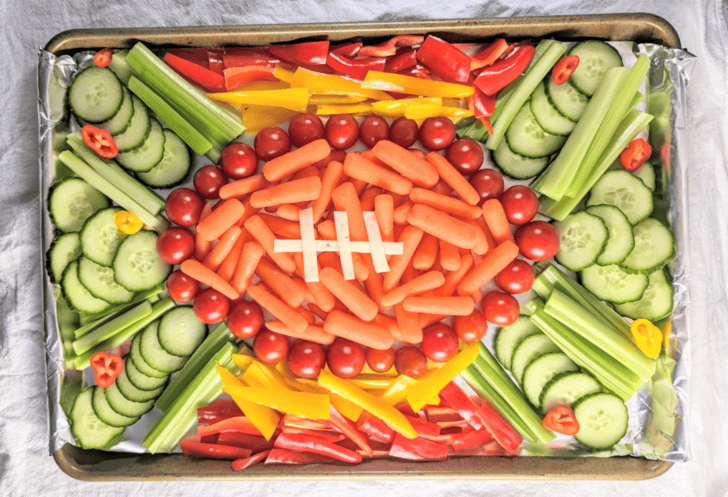 A vegetable platter arranged in the shape of a football on a tray. Carrots form the football, with celery sticks as stitches. Surrounding are sliced cucumbers, red and yellow bell peppers, cherry tomatoes, and celery sticks, all on foil.