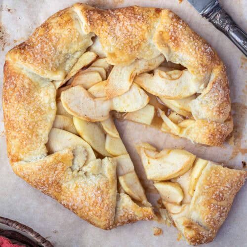 A rustic apple galette with flaky, golden-brown crust, sprinkled with sugar, is sliced on parchment paper. The filling showcases layered apple slices. A knife is partially visible at the top right corner.