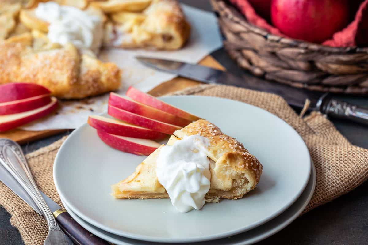 A slice of apple galette topped with whipped cream on a plate, with sliced red apples beside it. In the background, there's more tart on parchment paper and a basket of red apples. A knife rests nearby on a burlap cloth.