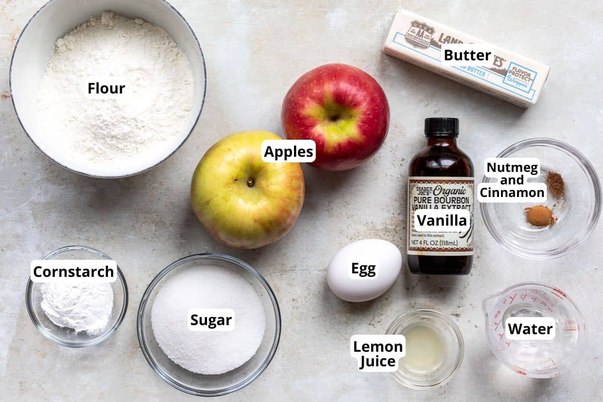 Top view of ingredients for baking on a light surface: a bowl of flour, apples, a stick of butter, a bowl of cornstarch, a bowl of sugar, an egg, vanilla extract, lemon juice, nutmeg and cinnamon mix, and water. Each is labeled.