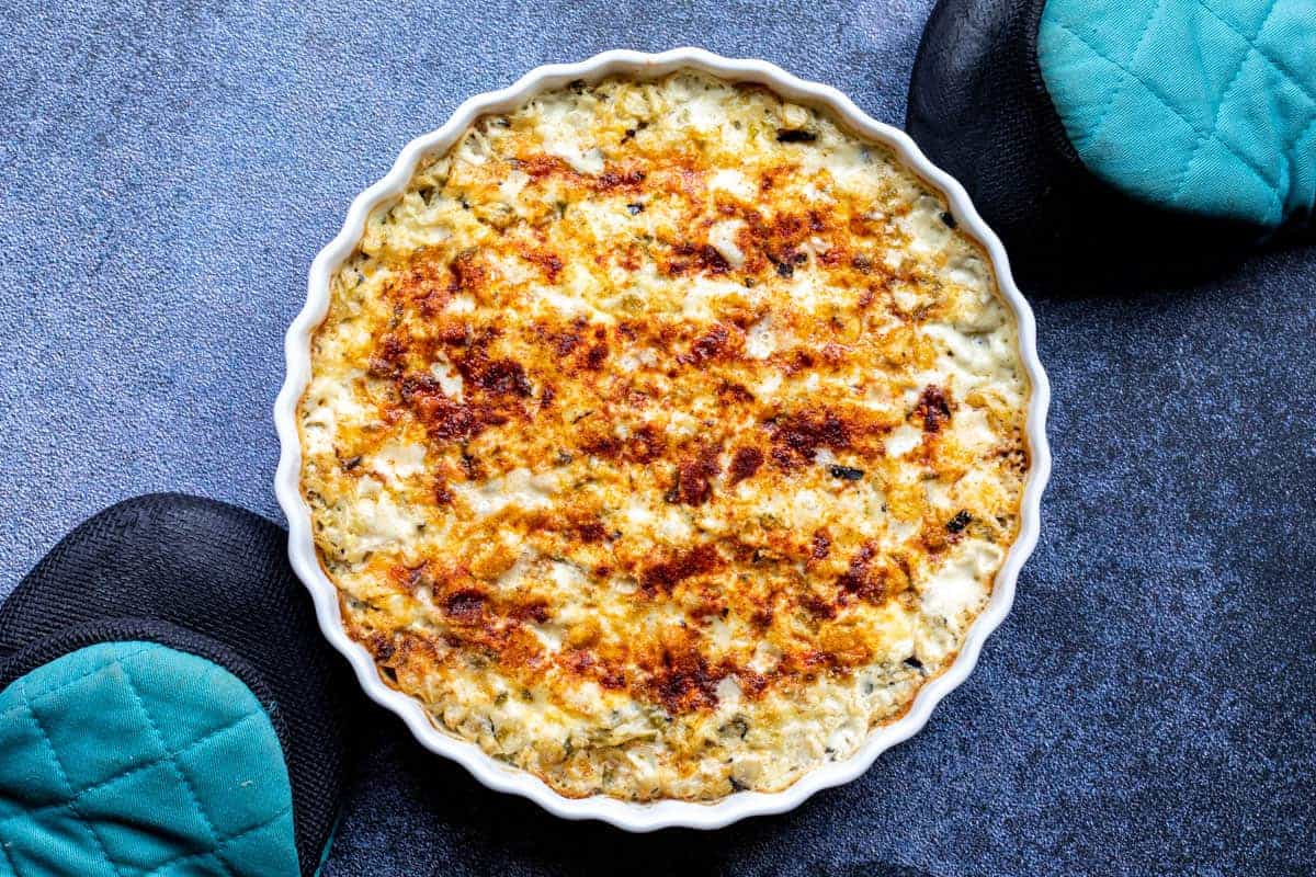 A baked casserole dish filled with a golden-brown crust, possibly breaded or topped with cheese. It is surrounded by two teal quilted oven mitts on a dark, textured surface.