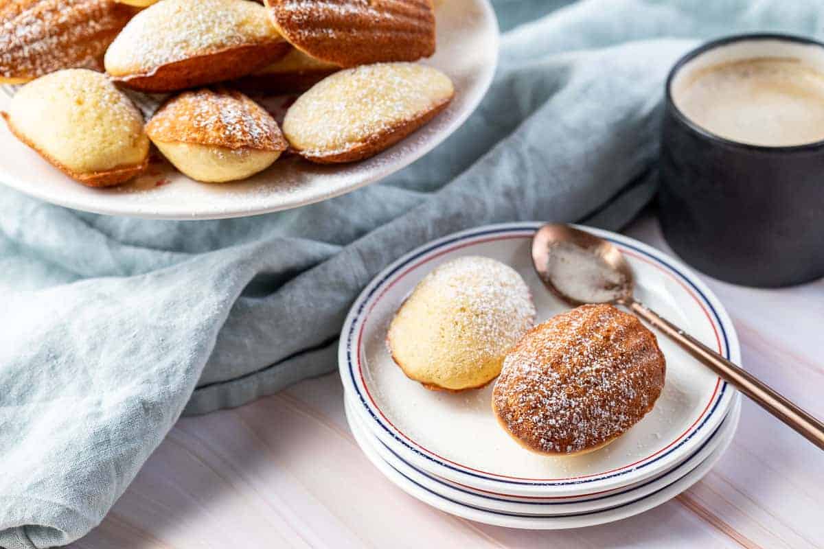 small plate with powdered sugar covered Madeleines next to cake stand with Madeleine cookies and cup of coffee.